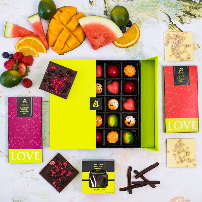 Half-open truffle box shows various colored truffles. Fruit displayed shows ingredients in candies. square pieces of dark chocolate raspberry inclusion bar and white chocolate passionfruit inclusion bar show texture and quality of chocolate.